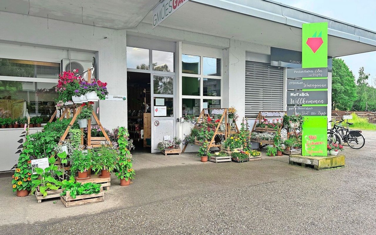 Im Eingangsbereich stehen Blumen und Setzlinge zum Verkauf. Trotz des Schildes erinnert der Hofladen an seine frühere Bestimmung als Garage. Von Vorteil ist die grosszügig bemessene Parkplatzzone.