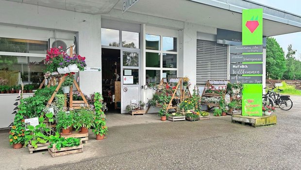 Im Eingangsbereich stehen Blumen und Setzlinge zum Verkauf. Trotz des Schildes erinnert der Hofladen an seine frühere Bestimmung als Garage. Von Vorteil ist die grosszügig bemessene Parkplatzzone.